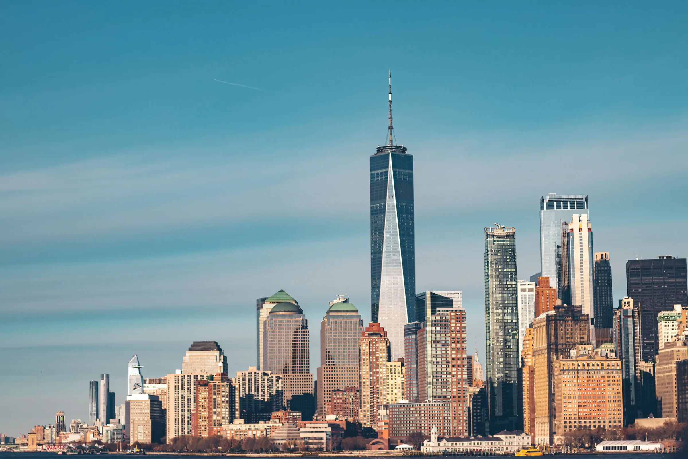 The Lower Manhattan skyline and One World Trade Center rising above the harbour under a clear blue sky.