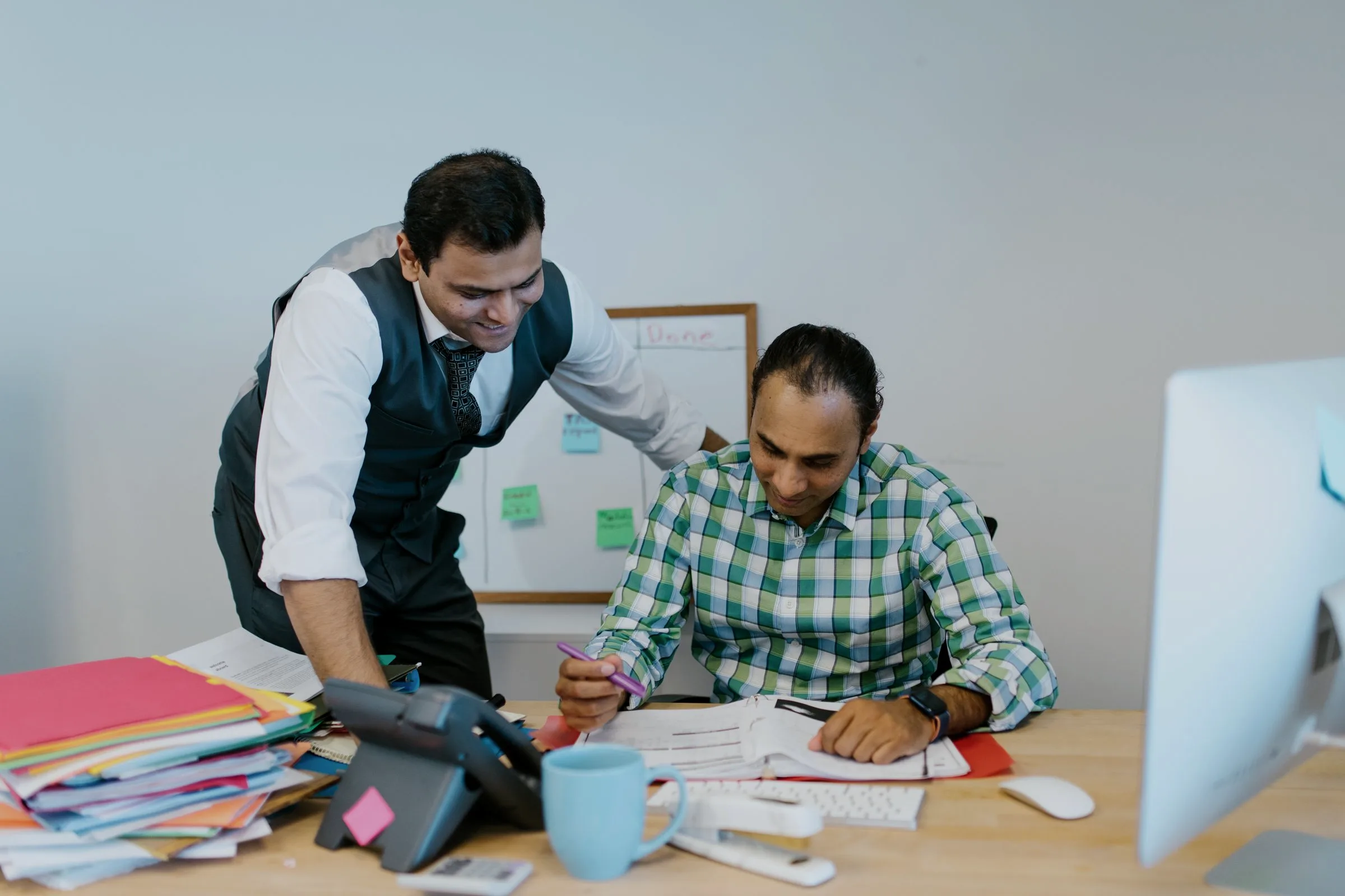 A colleague leaning over to review a printed page with a seated teammate at his desk.