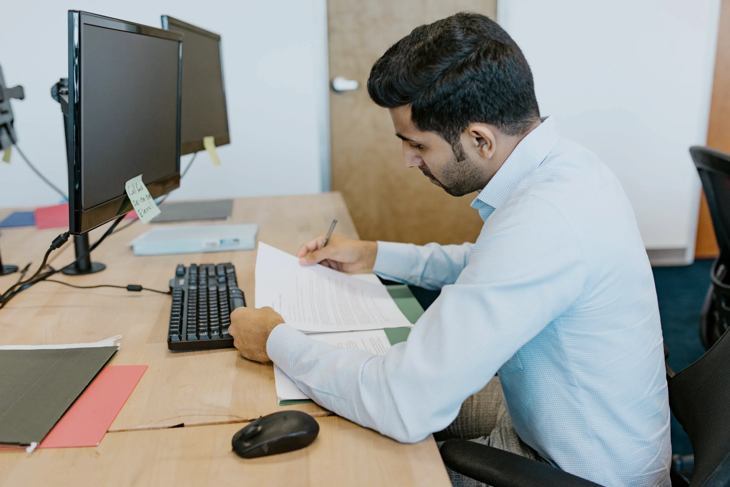 An accountant writing on a printed page at his desk, dual monitors and colour-coded folders in reach.